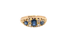 A yellow gold ring featuring a central oval-cut blue sapphire flanked by smaller round-cut blue sapphires and clear diamonds, set against a plain white background.