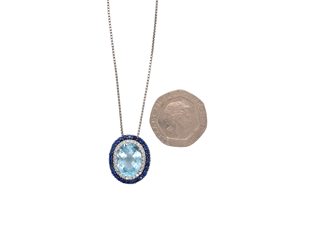 An oval pendant necklace with a light blue central stone and a halo of small dark blue stones, shown on a silver-toned chain next to a UK ten-pound coin for scale, on a plain white background.