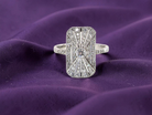 An Art Deco panel shaped cluster ring. The head measures 16x10mm, depth 3.4mm, and has two fan shaped designs that feature above and below a brilliant cut white diamond centre stone which measures 3.4mm
