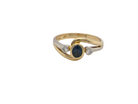An elegant 18 carat gold three-stone ring featuring a rich, oval-cut dark blue sapphire at the centre, measuring 5 × 4mm, secured in a classic rim (bezel) setting.