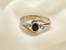 An elegant 18 carat gold three-stone ring featuring a rich, oval-cut dark blue sapphire at the centre, measuring 5 × 4mm, secured in a classic rim setting.