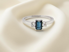 An elegant three-stone ring crafted in 18 carat white gold, featuring a striking rectangular dark blue sapphire at the centre, measuring 6 × 4 mm 