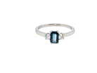 An elegant three-stone ring crafted in 18 carat white gold, featuring a striking rectangular dark blue sapphire at the centre, measuring 6 × 4 mm and weighing  0.50 carats.