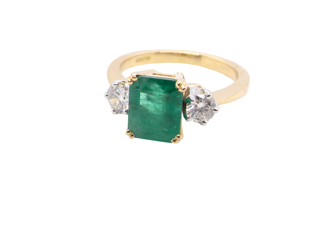 An exceptional three-stone ring featuring a vibrant step-cut emerald to the centre, measuring 8.6 x 7.0mm and weighing approximately 2.10 carats. The emerald displays a beautiful grass-green colour and is secured in a  four-claw setting.