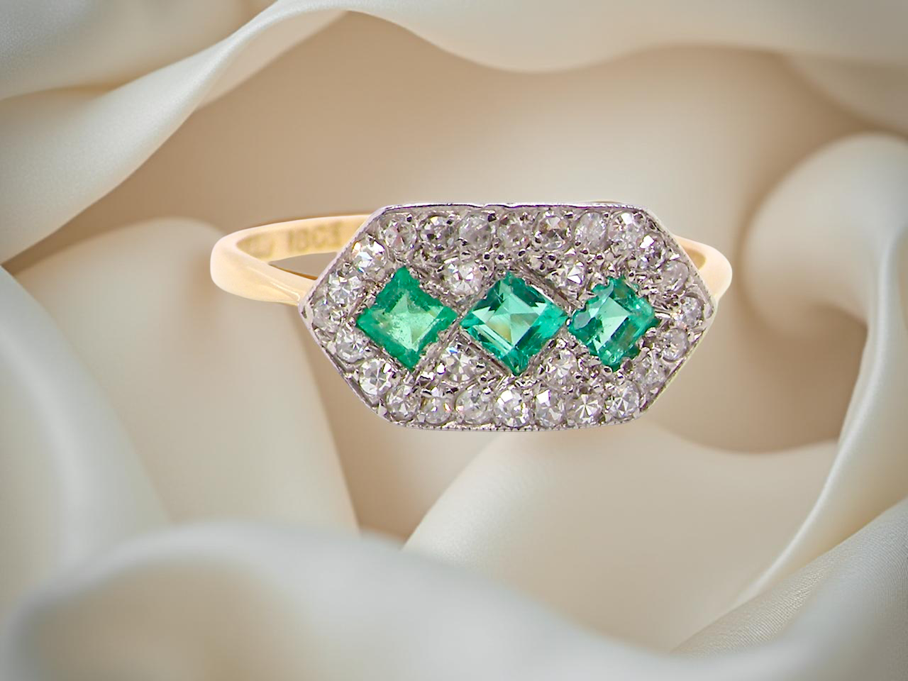 Edwardian era cluster ring, beautifully handcrafted with a trio of vibrant emeralds surrounded by a halo of antique old-cut diamonds. 