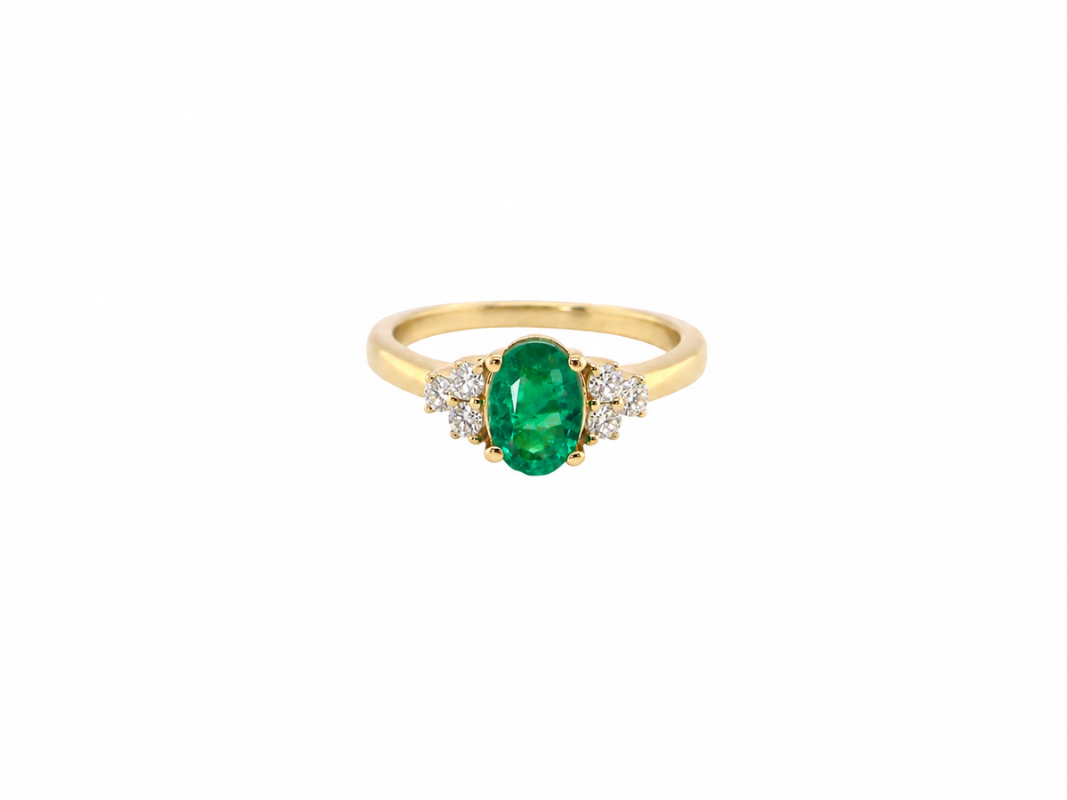 A yellow gold ring featuring an oval-cut green emerald center stone, flanked by small clusters of clear stones on either side, set against a plain white background.