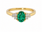 A yellow gold ring featuring an oval-cut green emerald center stone, flanked by small round clear stones on each side, set against a plain white background.
