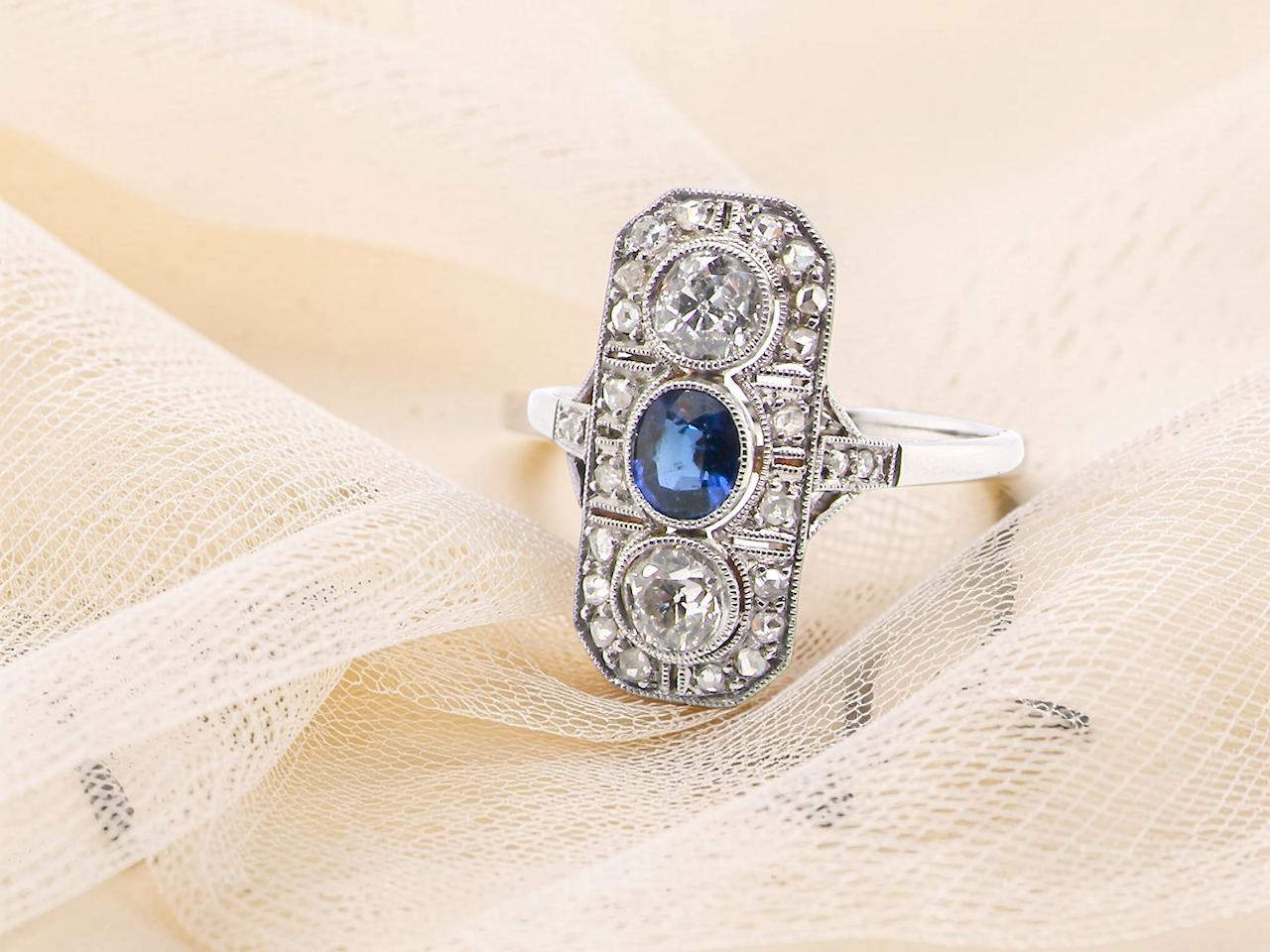 The plaque design has at its centre a mid to dark blue sapphire, 5x4mm (est 0.40 carats) mille grain set. Above and below to Old European Cut white diamonds. 