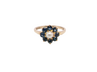 A vintage-style gold ring featuring a central white pearl surrounded by a halo of dark blue gemstones, set against a plain white background.
