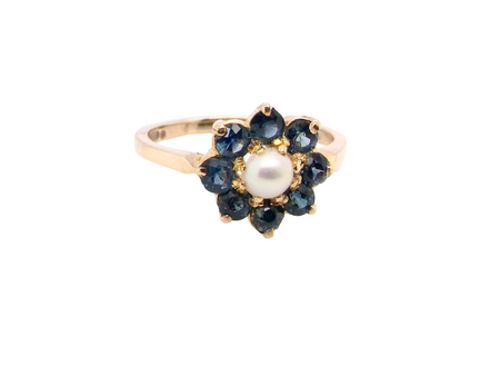 A yellow gold ring featuring a flower-shaped cluster with a central white pearl surrounded by dark blue round-cut gemstones, set against a plain white background.