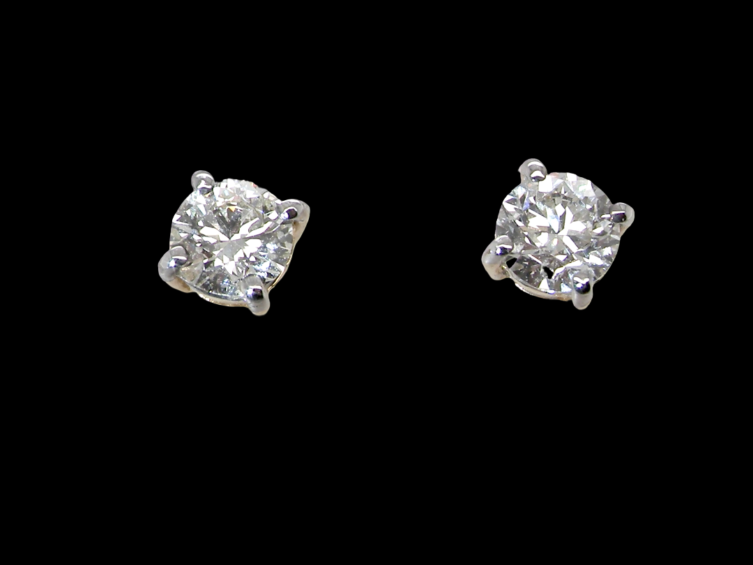 A pair of classic solitaire stud earrings featuring round brilliant-cut diamonds in four-prong silver settings, presented on a solid black background.