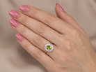A close-up of a hand with pink manicured nails wearing a gold ring featuring a green peridot stone surrounded by a halo of small clear stones, set against a soft beige fabric background.