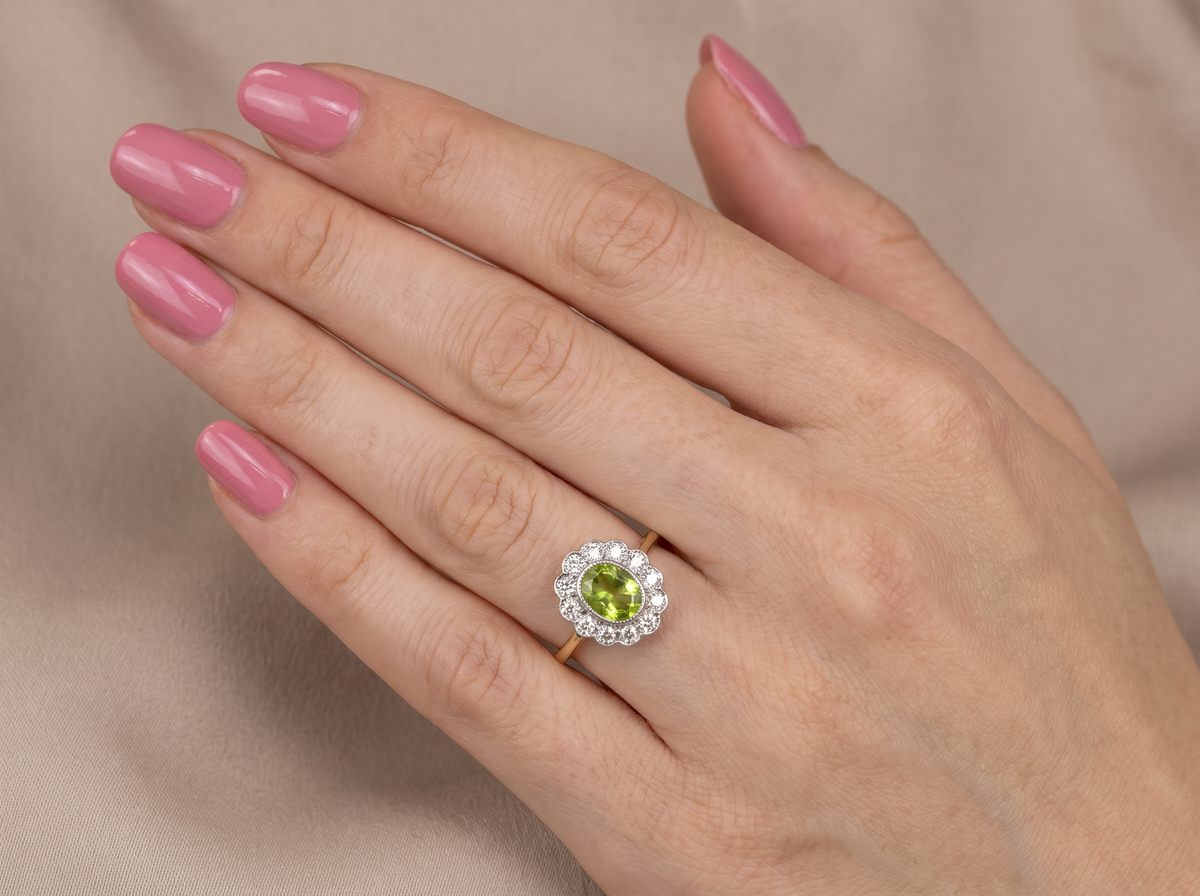 A close-up of a hand with pink manicured nails wearing a gold ring featuring a green peridot stone surrounded by a halo of small clear stones, set against a soft beige fabric background.