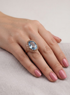 A close-up of a hand with pink manicured nails wearing a gold ring featuring an oval light blue gemstone in an openwork filigree setting, resting on a textured light gray fabric surface.