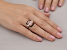 A close-up of a hand wearing a ring with a rectangular pale pink gemstone, a black inlay, and a rose gold band, set against a neutral gray background.