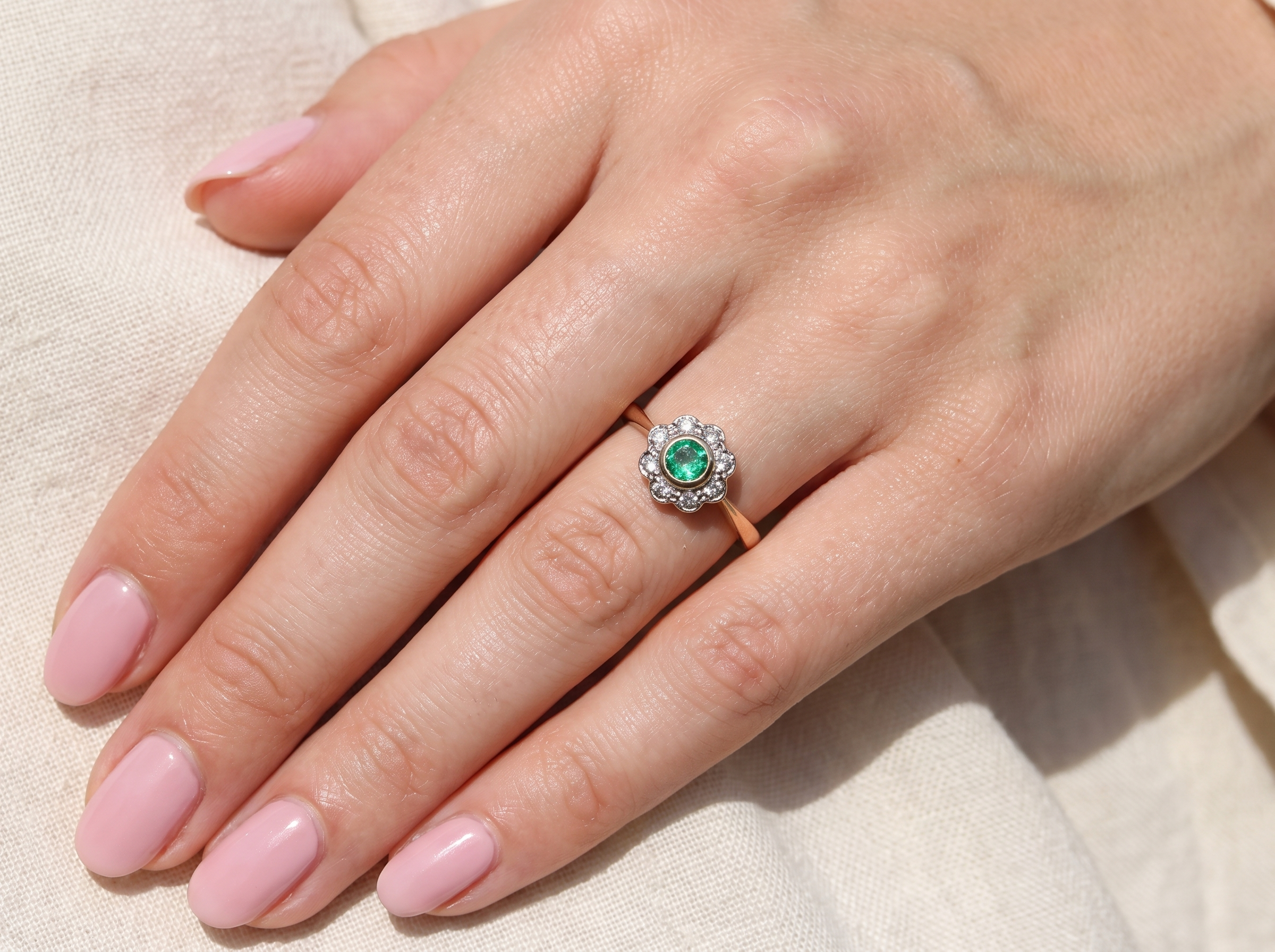 A close-up of a hand wearing a ring with a round green center stone surrounded by small clear stones, set on a rose gold band, resting on a light beige fabric background.