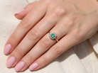 A close-up of a hand wearing a ring with a round green center stone surrounded by small clear stones, set on a rose gold band, resting on a light beige fabric background.