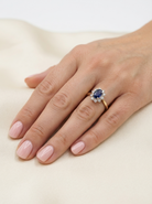 A close-up of a hand with light pink manicured nails wearing a gold ring featuring a dark blue oval center stone surrounded by small clear stones, resting on a soft cream fabric background.