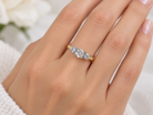 A close-up of a hand wearing a yellow gold three-stone ring with clear round-cut diamonds. The hand is shown against a soft, out-of-focus white background with a hint of green foliage.