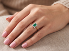 A close-up of a hand wearing a gold ring with a rectangular green emerald-cut center stone and small clear accent stones, resting on a beige textured fabric surface.