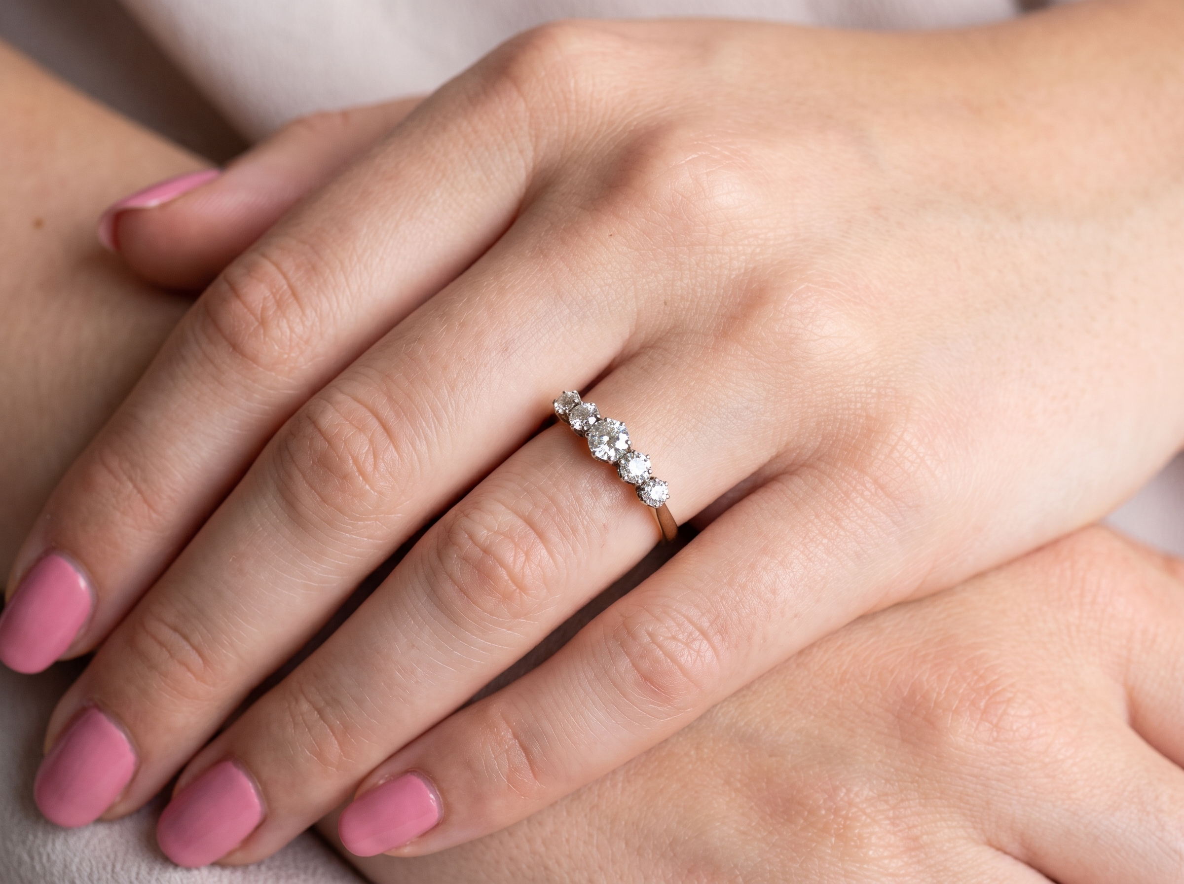 A close-up of a hand with pink manicured nails wearing a delicate gold ring set with small clear stones, resting on another hand against a soft neutral background.
