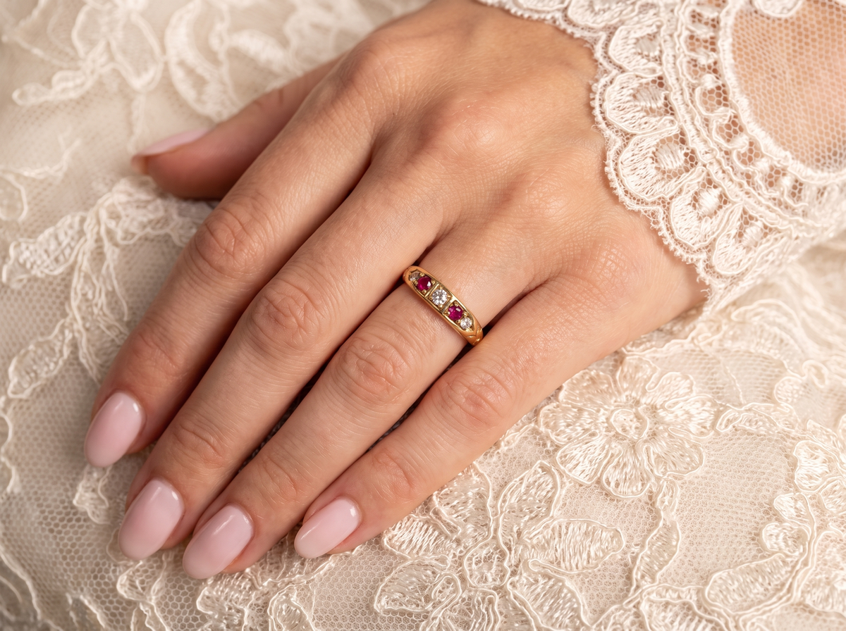 A close-up of a hand wearing a delicate gold band ring with small red and clear stones, resting on cream lace fabric.