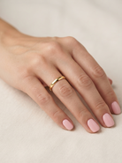 A close-up of a hand with light pink manicured nails wearing a simple, polished gold band ring on the ring finger, resting on a soft beige fabric background.