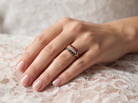 A close-up of a hand wearing a gold ring with rows of small pink, blue, and clear stones, resting on white lace fabric.