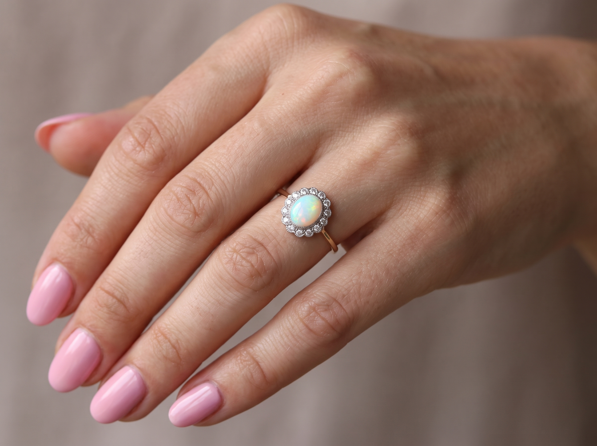 A close-up of a hand wearing a gold ring with a round iridescent opal center stone and a halo of small clear stones, set against a soft neutral background.