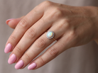 A close-up of a hand wearing a gold ring with a round iridescent opal center stone and a halo of small clear stones, set against a soft neutral background.