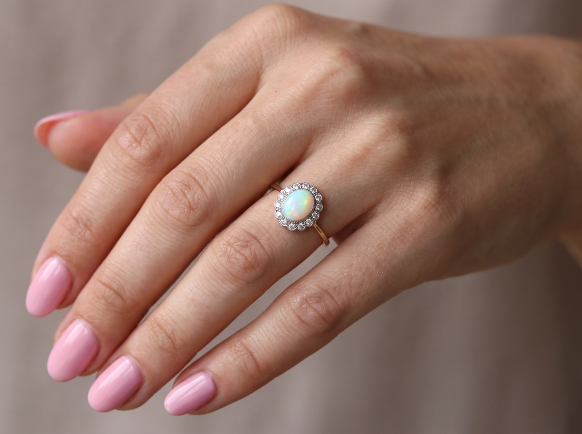 A close-up of a hand wearing a gold ring with a round iridescent opal center stone and a halo of small clear stones, set against a soft neutral background.