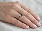 A close-up of a hand wearing a delicate ring with a gold leaf design, a clear center stone, and small dark blue accent stones, resting on a soft beige fabric background.
