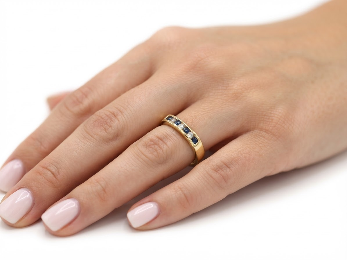 A close-up of a hand wearing a yellow gold eternity ring set with small round diamonds and dark blue round gemstones, shown against a plain white background.