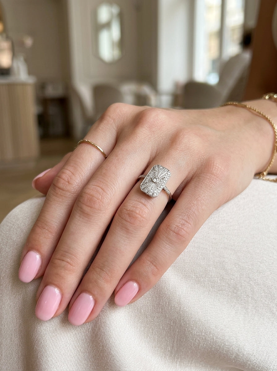 hand An Art Deco panel shaped cluster ring. The head measures 16x10mm, depth 3.4mm, and has two fan shaped designs that feature above and below a brilliant cut white diamond centre stone which measures 3.4mm (0.15 carats est)
