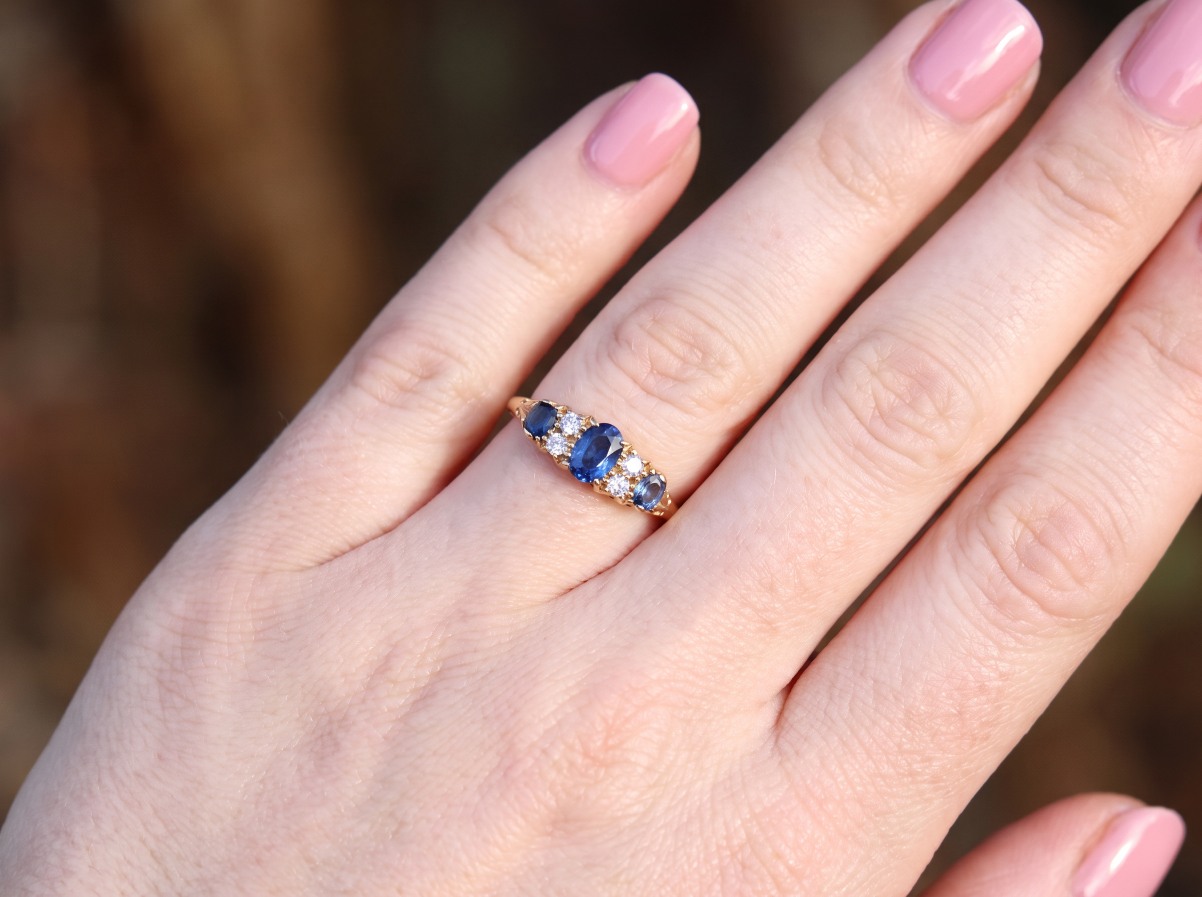 A close-up of a hand wearing a yellow gold ring with a central oval blue sapphire and smaller round blue and clear stones on the side, set against a blurred natural background.