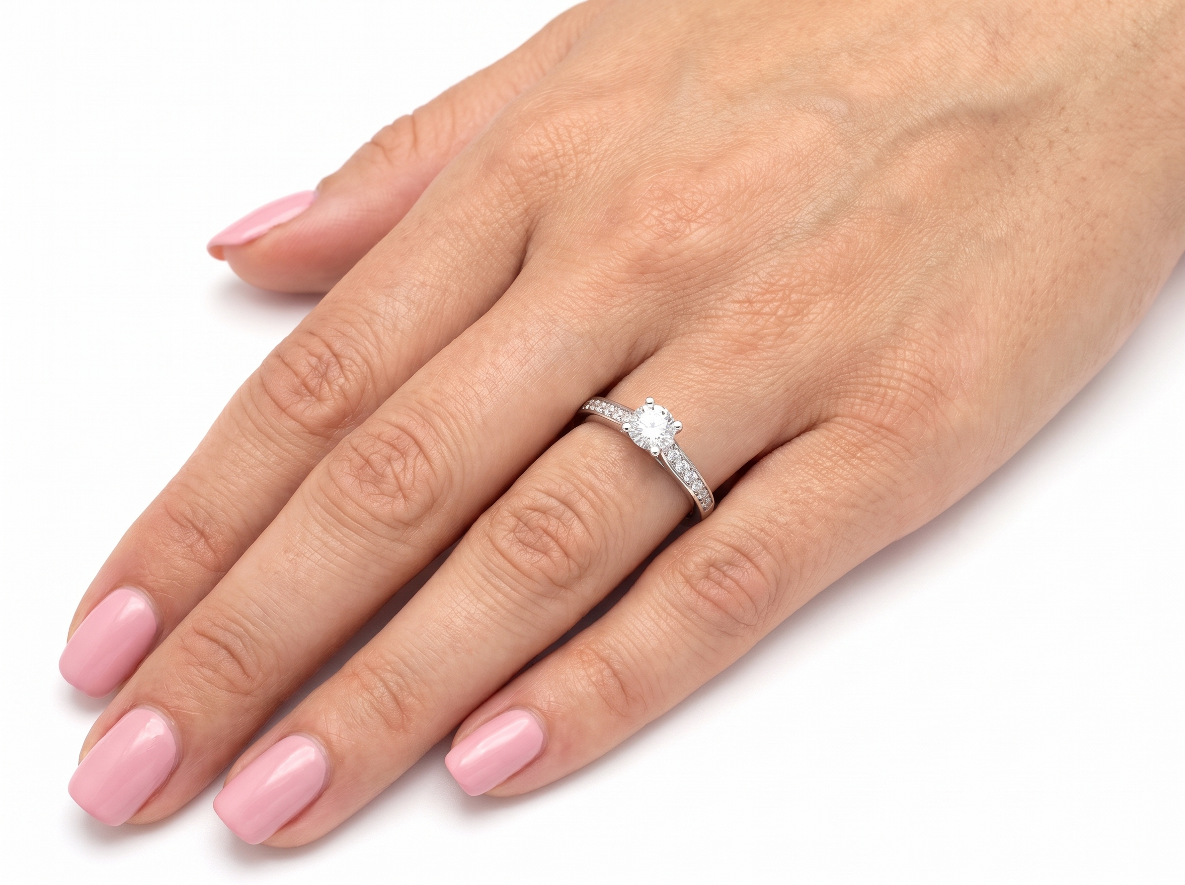A close-up of a hand with pink manicured nails wearing a diamond engagement ring on the ring finger, set against a plain white background.