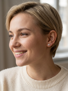 A close-up portrait of a smiling woman with short blonde hair wearing a small square silver stud earring, set against a softly blurred indoor background.