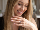 A close-up lifestyle shot of a smiling blonde woman wearing a silver band ring with a clear center stone on her ring finger, with her hand raised in front of her face.