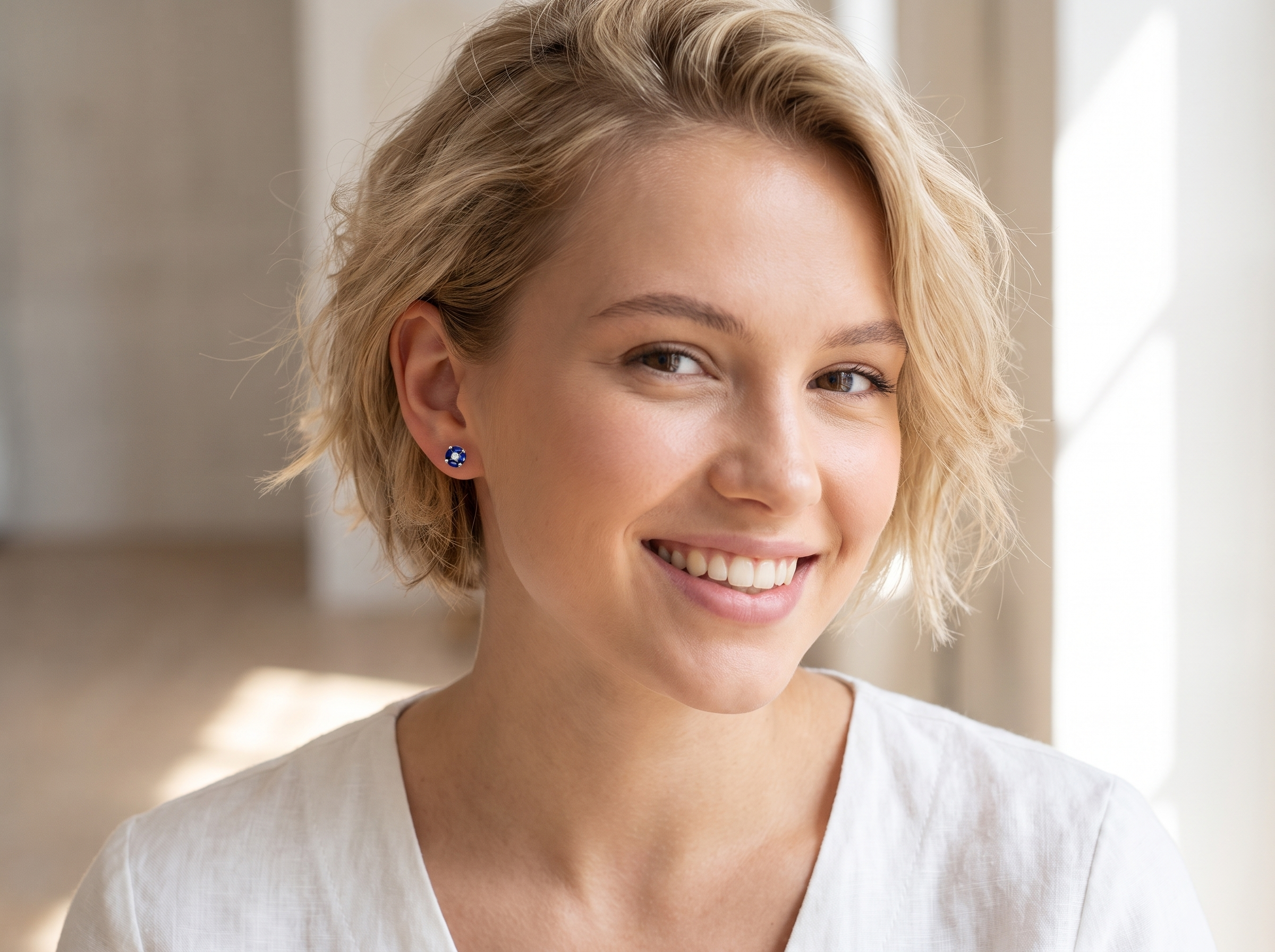 A smiling blonde woman wearing a small blue stud earring, shown in a bright indoor setting with soft natural light.