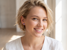A smiling blonde woman wearing a small blue stud earring, shown in a bright indoor setting with soft natural light.