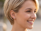 A close-up side profile of a smiling woman with short blonde hair wearing a small round stud earring.