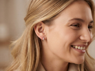 A close-up side profile of a smiling blonde woman wearing a small hoop earring with a pink stone and a tiny clear accent stone.
