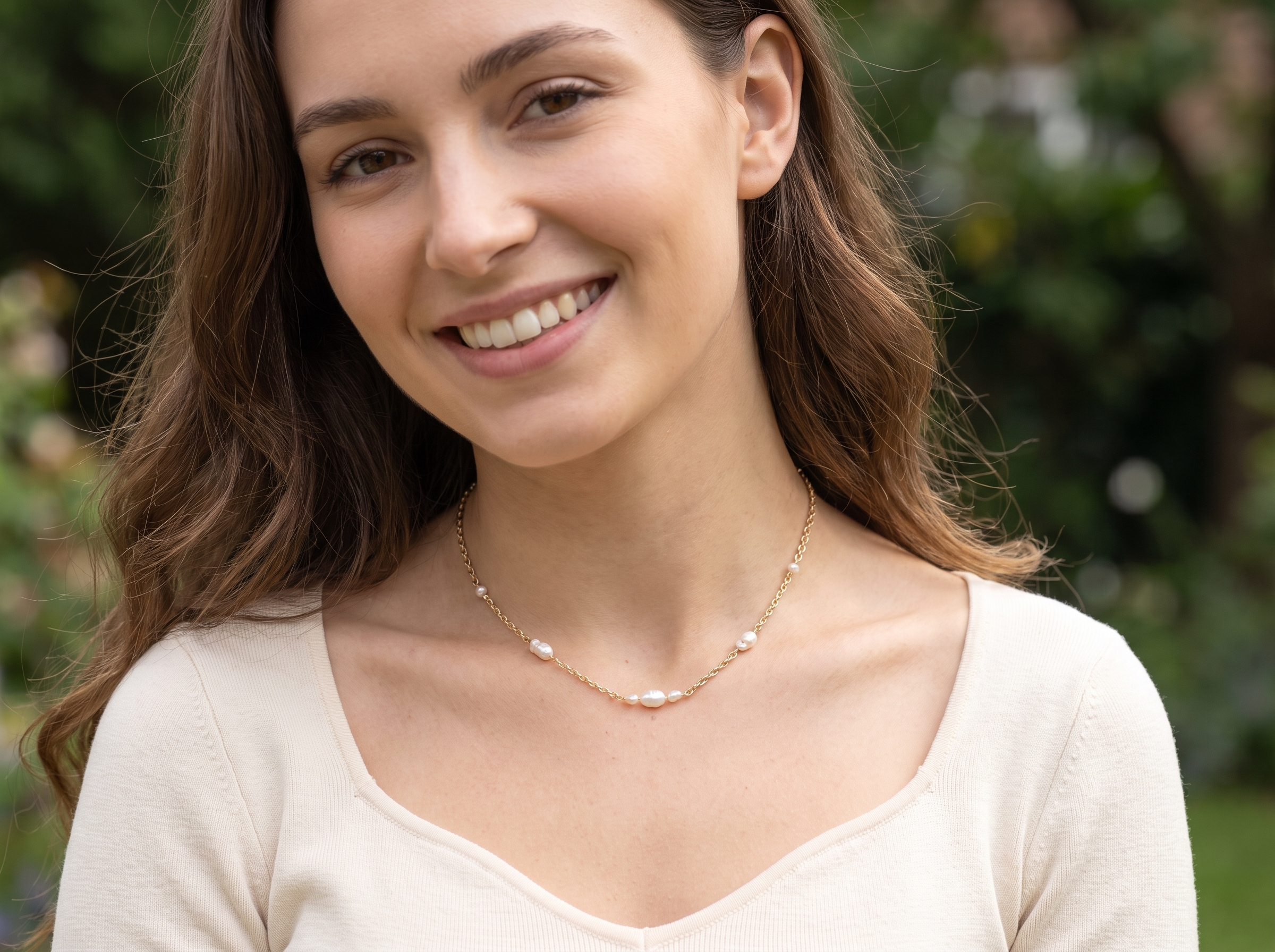 A smiling woman with long brown hair wearing a delicate gold chain necklace with small white pearl-like beads, photographed outdoors against a blurred green background.