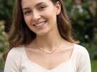 A smiling woman with long brown hair wearing a delicate gold chain necklace with small white pearl-like beads, photographed outdoors against a blurred green background.