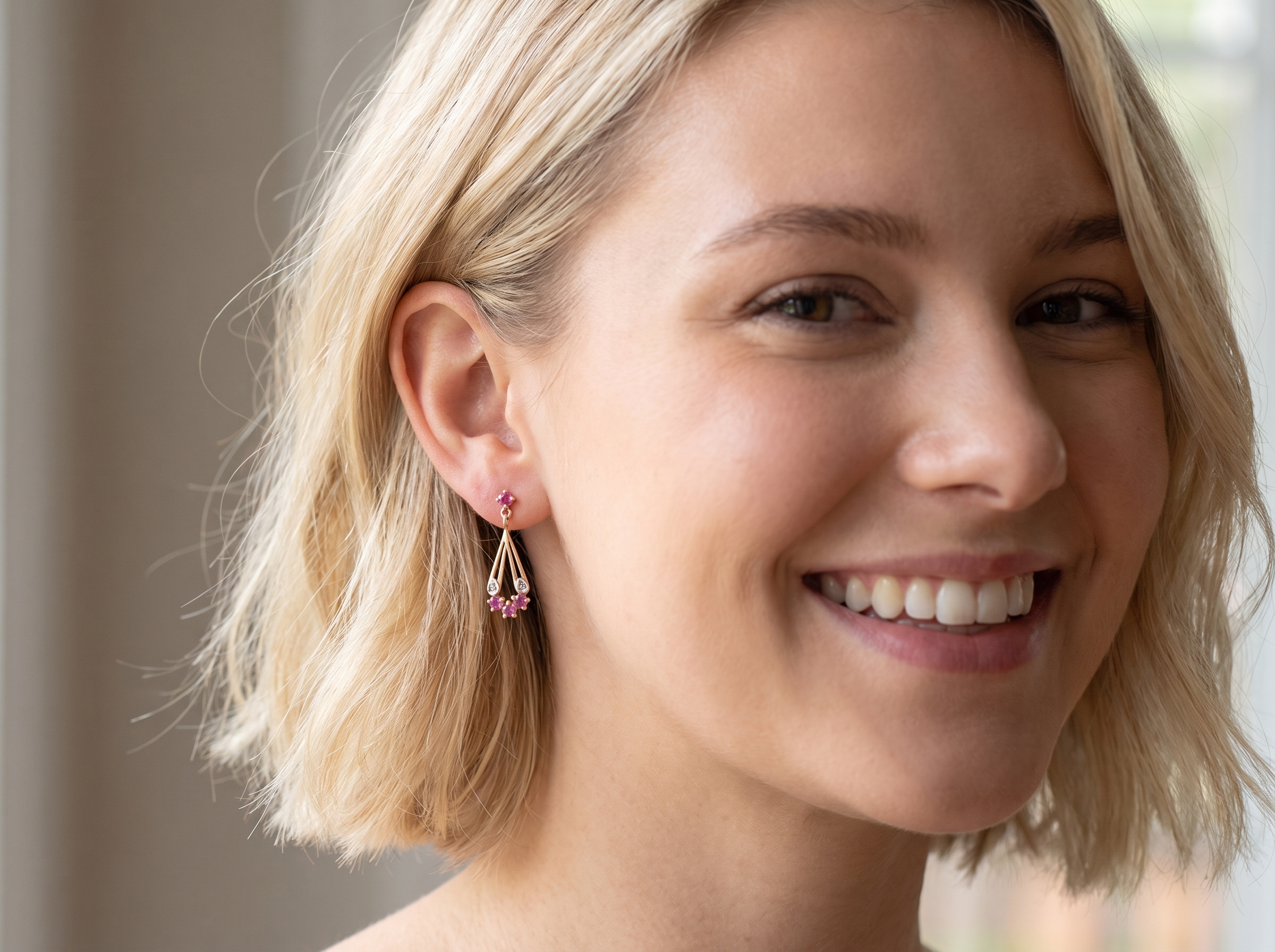 A close-up of a smiling blonde woman wearing a delicate gold dangle earring with small pink gemstones, set against a softly blurred indoor background.