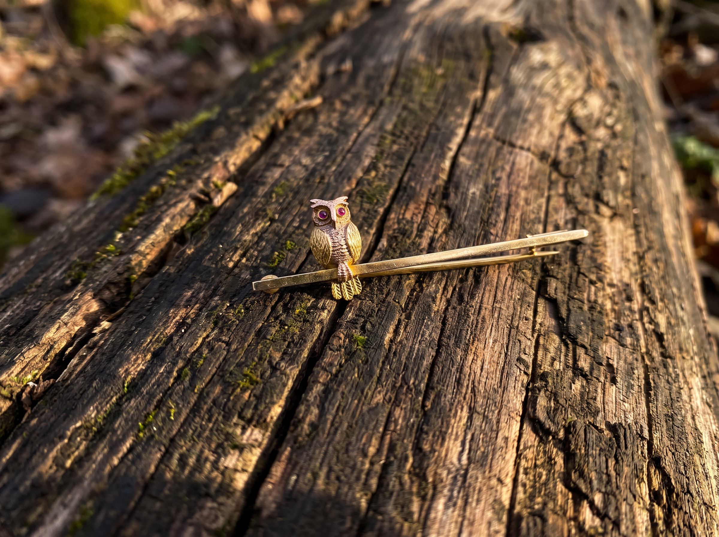 A small owl-shaped hair pin or hair accessory with a metallic finish rests on a weathered log outdoors.