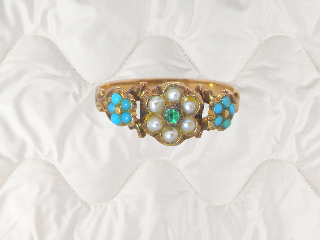 An antique Victorian gold ring with a floral design featuring white pearls, a central green emerald, and turquoise blue stones, displayed on a white quilted fabric background.