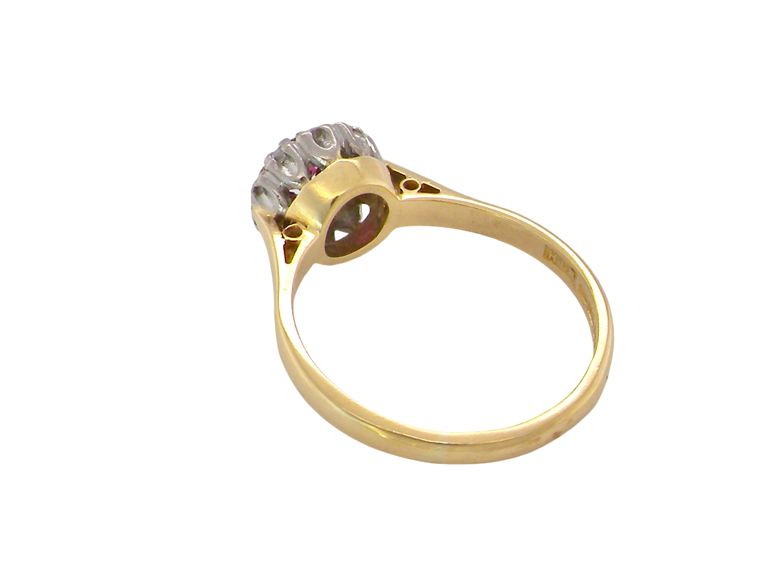 A yellow gold ring with a floral cluster setting featuring clear gemstones and a tiny red accent stone, shown on a plain white background.