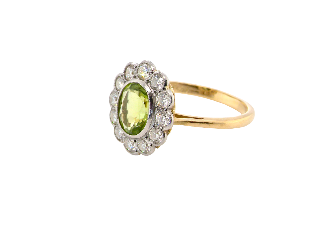 A yellow gold ring featuring an oval green peridot stone surrounded by a halo of small clear stones, set on a plain white background.