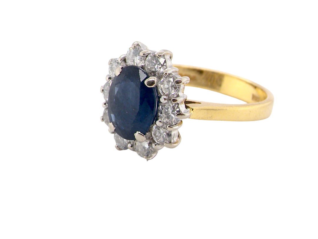 A yellow gold ring featuring an oval dark blue sapphire center stone surrounded by a halo of small clear stones, set on a plain white background.